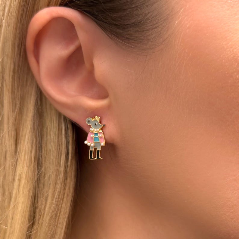 May include: Gold-tone mouse stud earrings. The earrings feature a gray mouse wearing a pink cape with white trim, a blue shirt, and black boots. The mouse has a gold crown. The earrings are set against a blurred background.
