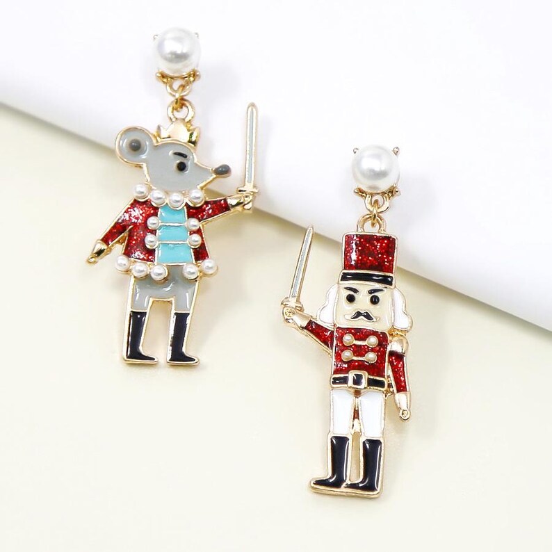May include: A pair of festive dangle earrings featuring a mouse and a nutcracker design. The mouse is grey with a red jacket and a crown, while the nutcracker wears a red uniform. Both hold swords and are accented with pearls and gold-tone metal.
