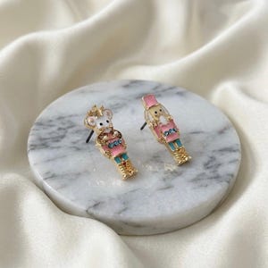 May include: A pair of stud earrings featuring a gold-tone metal nutcracker and a mouse nutcracker design. Both figures are decorated with pink and blue enamel, small clear rhinestones, and gold-tone accents. The earrings have a post-back closure.