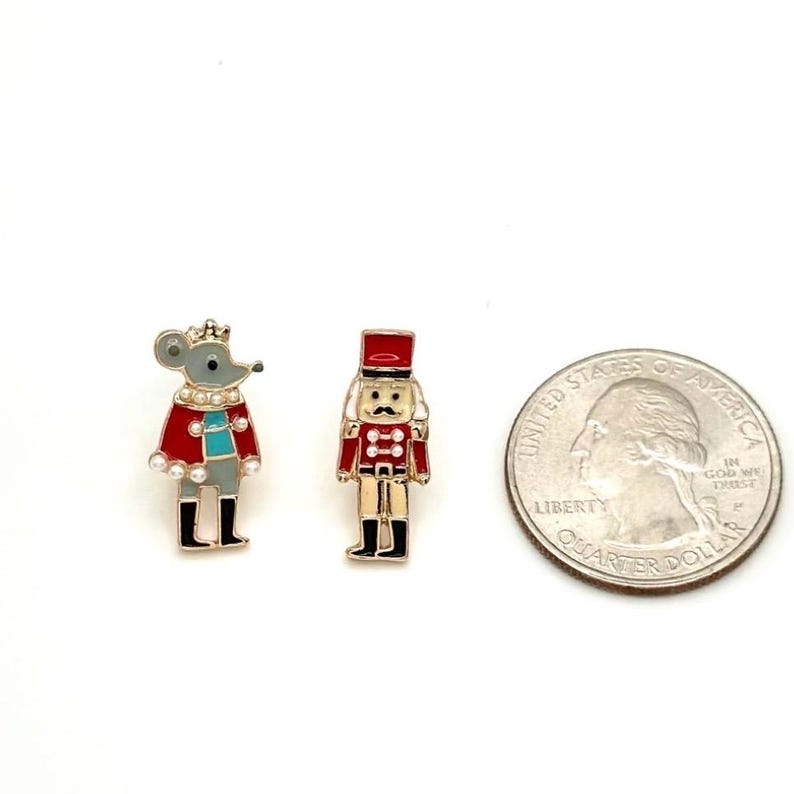 May include: Two Christmas-themed enamel pins: a mouse king in a red coat and a nutcracker soldier in a red uniform. A U.S. quarter coin is included for size comparison. The pins have gold-colored metal accents and black boots.
