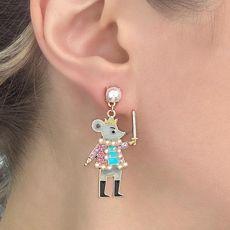 May include: A gold-coloured earring depicting a mouse character wearing a crown, pink jacket, and holding a sword. The mouse has a grey body, black boots, and a turquoise chest. A pearl tops the earring.