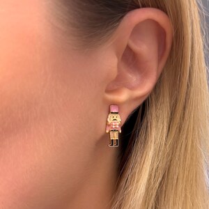 May include: Gold-toned stud earrings featuring a pink and white nutcracker soldier design. The earrings have a detailed, colorful enamel finish, with a small, rectangular shape. The earrings are shown on a person's ear.