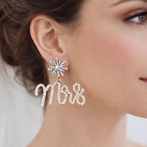 May include: Sparkling "Mrs" earrings. The earrings feature a snowflake-shaped stud with a central crystal and a dangling "Mrs" script, all encrusted with clear crystals. A glamorous accessory for a bride.