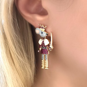 May include: A single, whimsical earring featuring a mouse soldier design. The mouse wears a crown, a white cape, and a burgundy uniform with gold accents. It holds a sword, with silver and gold details.