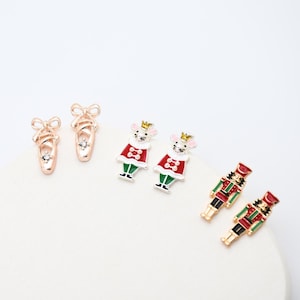 May include: Three pairs of stud earrings featuring rose gold ballet slippers with clear stones, white mice wearing red and green outfits with gold crowns, and red, green, and gold nutcracker figures with black accents, all crafted with enamel details.