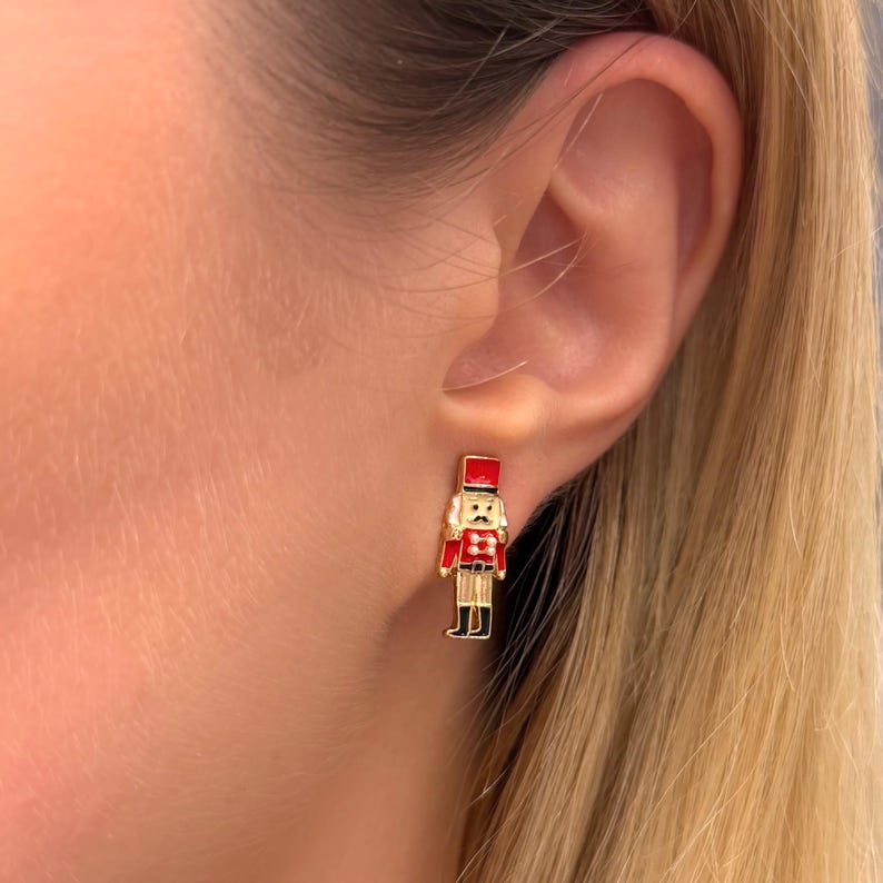 May include: Gold-toned stud earrings featuring a miniature, detailed design of a toy soldier. The soldier is dressed in a red uniform with black boots and a red hat. The earrings are a festive accessory.