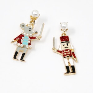 May include: A pair of gold-coloured earrings featuring a mouse and a nutcracker design. The mouse is grey with a red jacket and a crown, while the nutcracker is red and white. Both hold swords and are accented with pearls.