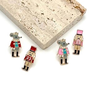 May include: Four decorative enamel pins featuring a mouse and a nutcracker soldier design. The mouse pins have gray bodies, crowns, and capes in red or pink. The nutcracker pins are in red and pink with black boots.