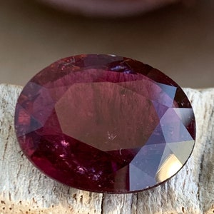 May include: A deep purple gemstone with a faceted cut. The gemstone is oval shaped and has a high polish.