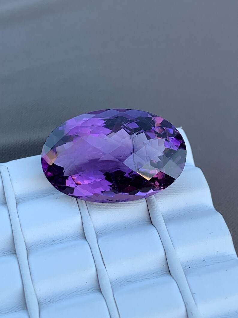 K&ouml;nnte beinhalten: Ein gro&szlig;er, ovaler Amethyst-Edelstein mit tiefem Lilafarbton. Der Edelstein ist facettiert und hat einen hohen Glanz.