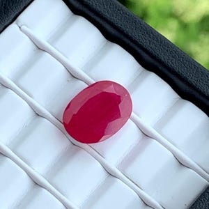 Peut inclure: Une pierre précieuse ovale rouge vif avec une surface lisse et polie. La pierre précieuse est présentée sur un fond blanc.