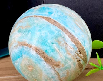 May include: A polished sphere of blue and tan stone. The stone has a marbled appearance with swirling patterns of light blue, tan, and cream colors. The sphere is smooth and round, with a natural, earthy aesthetic.