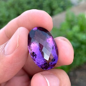 K&ouml;nnte beinhalten: Ein gro&szlig;er, ovaler Amethyst-Edelstein mit tiefem Lilafarbton. Der Edelstein ist facettiert und hat einen hohen Glanz.