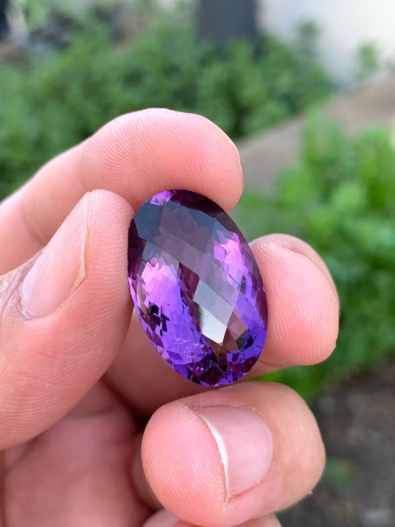 K&ouml;nnte beinhalten: Ein gro&szlig;er, ovaler Amethyst-Edelstein mit tiefem Lilafarbton. Der Edelstein ist facettiert und hat einen hohen Glanz.