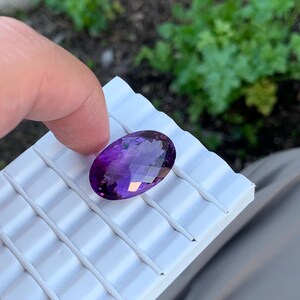 K&ouml;nnte beinhalten: Ein gro&szlig;er, ovaler Amethyst-Edelstein mit tiefem Purpurton. Der Edelstein ist facettiert und hat eine hohe Klarheit. Er wird auf einer wei&szlig;en, strukturierten Oberfl&auml;che pr&auml;sentiert.