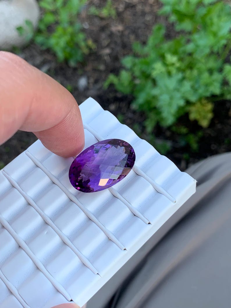 K&ouml;nnte beinhalten: Ein gro&szlig;er, ovaler Amethyst-Edelstein mit tiefem Purpurton. Der Edelstein ist facettiert und hat eine hohe Klarheit. Er wird auf einer wei&szlig;en, strukturierten Oberfl&auml;che pr&auml;sentiert.