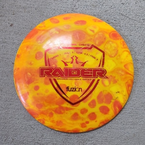 May include: A yellow and orange disc golf disc with the word "RAIDER" in red. The disc has the text "MAXIMUM DISTANCE DRIVER" and "fuzion". The disc also has the text "SPEED: 1 | GLIDE: 5 | TURN: -0.5 | FADE: 3".