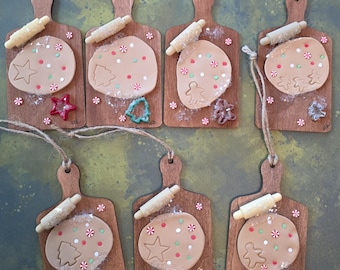 May include: Wooden ornaments shaped like cutting boards, each featuring a circle of dough with a star, gingerbread man, or Christmas tree cutout. Rolling pins and colorful sprinkles add detail. The ornaments are strung with twine.