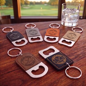 May include: Six metal bottle opener keychains with leather-like rectangular patches in black, gray, orange, tan, and brown. Each patch features a circular logo with crossed golf clubs and the text Cedar Ridge Classic 2027. A glass of water.