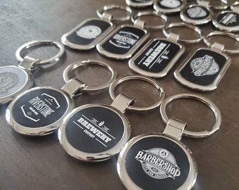 May include: A collection of silver-toned metal keychains featuring circular and rectangular shapes with black inserts. Each keychain displays white printed logos and text, including Riverstone, Brewery Company, and Barbershop, attached to split rings.