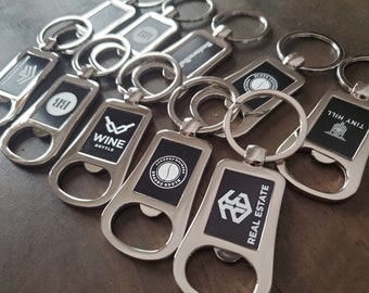 May include: A collection of silver-toned metal keychains, each featuring a bottle opener function and a rectangular black inlay. The inlays display various white printed logos and text, including "WINE BOTTLE," "BLACK COFFEE," and "REAL ESTATE."