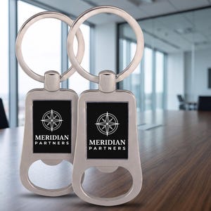 May include: Two silver-toned metal keychains featuring a rectangular black insert with a white compass rose graphic and the text Meridian Partners printed above the word Partners. Each keychain includes a circular ring and a bottle opener base.