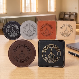 May include: A collection of six coasters in round and square shapes, featuring various colors including black, gray, brown, and tan. Each coaster is embossed with a circular logo depicting a church building and the text Grace Harbor Fellowship.