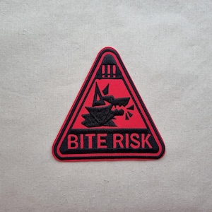 May include: A red triangular patch with a black border and a black illustration of a snarling wolf. The text "BITE RISK" is in black letters below the illustration. The patch is on a light beige fabric.