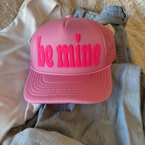 Puede incluir: Gorra rosa con las palabras "be mine" en fuente de burbujas y un pequeño corazón encima de la "i". La gorra tiene una parte trasera de malla y una visera curva. La gorra está sobre unos vaqueros azules.