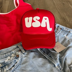 May include: Red trucker hat with a white "USA" logo in a retro font with a small red heart in the "A". The hat is on top of a pair of light blue denim shorts.
