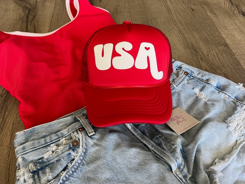 May include: Red trucker hat with a white "USA" logo in a retro font with a small red heart in the "A". The hat is on top of a pair of light blue denim shorts.