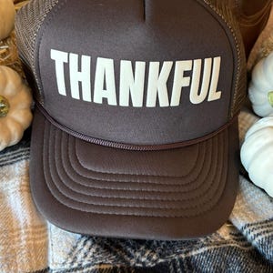 Op de afbeelding: Een bruine truckercap met een wit mesh achterpaneel. De voorkant van de cap heeft het woord "THANKFUL" in witte letters.