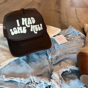 May include: A brown trucker hat with a white mesh back. The hat has white text that reads "I had some help" in a retro font.