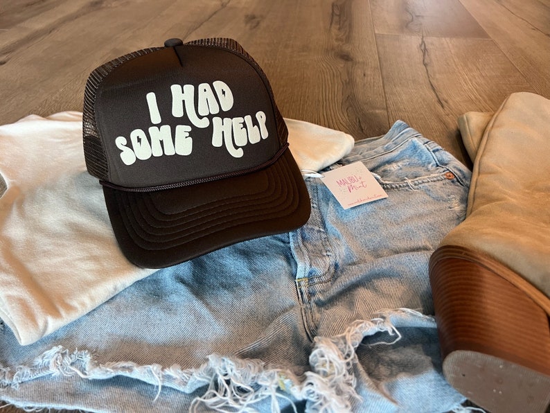 May include: A brown trucker hat with a white mesh back. The hat has white text that reads "I had some help" in a retro font.
