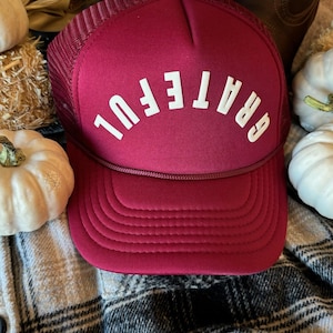 May include: A burgundy trucker hat with a white mesh back and the word "GRATEFUL" in white letters on the front.