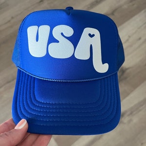 May include: A royal blue trucker hat with a white "USA" logo in a retro font. The "A" has a small white heart in the center.