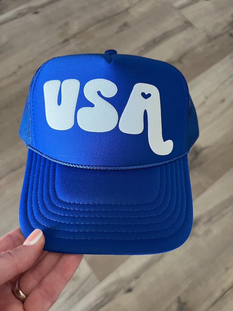 May include: A royal blue trucker hat with a white "USA" logo in a retro font. The "A" has a small white heart in the center.