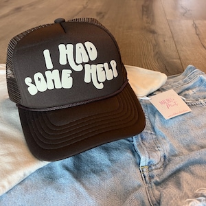 May include: A brown trucker hat with a white mesh back. The hat has white text that reads "I had some help" in a retro font.