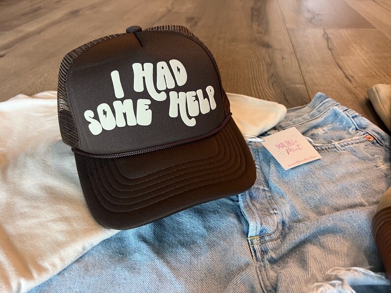 May include: A brown trucker hat with a white mesh back. The hat has white text that reads "I had some help" in a retro font.
