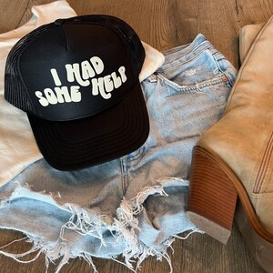 May include: A black trucker hat with a white logo that says "I had some help". The hat is on top of a pair of light blue denim shorts and a white tank top. A pair of tan suede boots are also in the image.