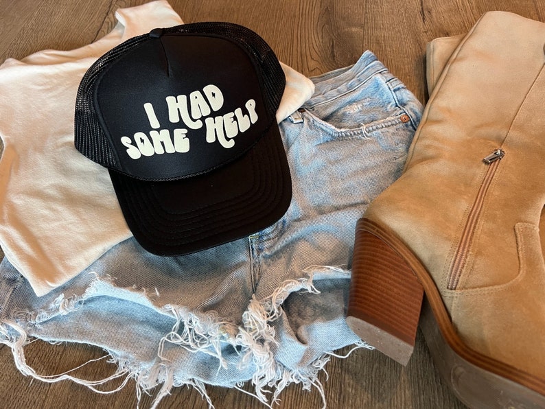 May include: A black trucker hat with a white logo that says "I had some help". The hat is on top of a pair of light blue denim shorts and a white tank top. A pair of tan suede boots are also in the image.