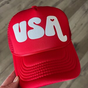 May include: Red trucker hat with a white "USA" logo in a retro font. The "A" has a small red heart on it.