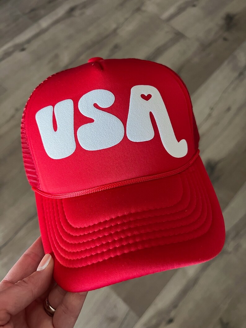 May include: Red trucker hat with a white "USA" logo in a retro font. The "A" has a small red heart on it.