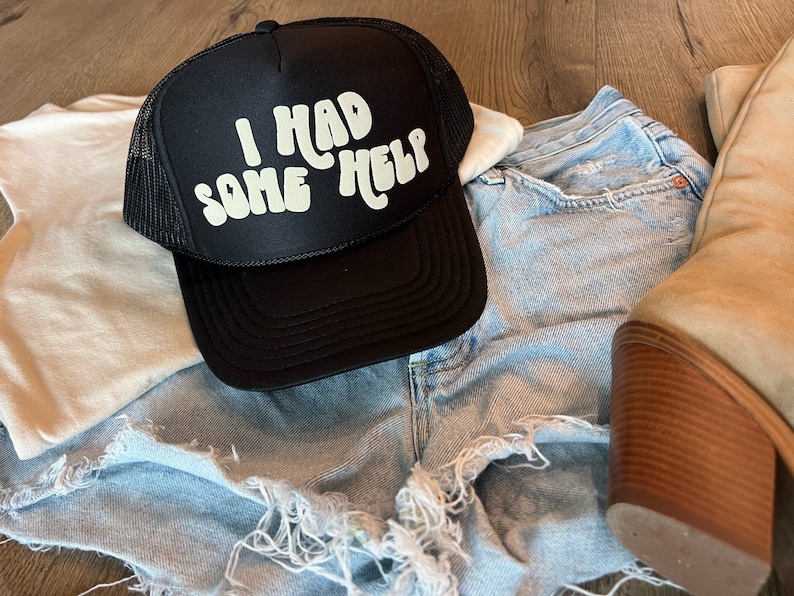 May include: A black trucker hat with a white mesh back. The hat has white text that reads "I had some help" in a retro font.