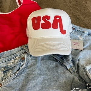 May include: White trucker hat with a red "USA" graphic and a small red heart. The hat is on a denim background.