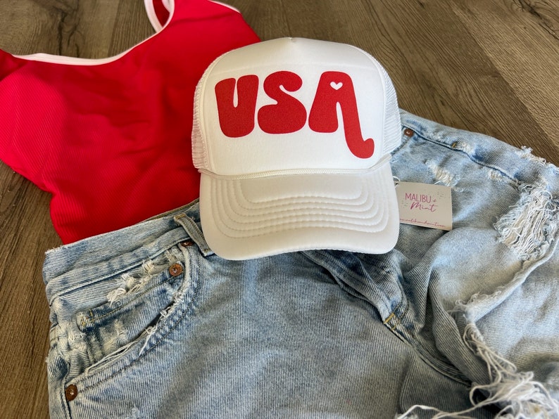 May include: White trucker hat with a red "USA" graphic and a small red heart. The hat is on a denim background.