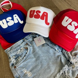 May include: Three trucker hats with a white "USA" graphic with a heart in the "A". The hats are blue, white, and red.