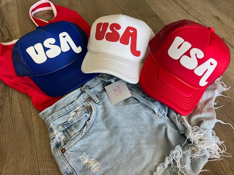 May include: Three trucker hats with a white "USA" graphic with a heart in the "A". The hats are blue, white, and red.