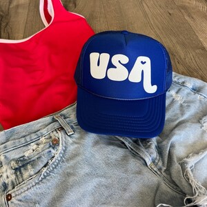 May include: A blue trucker hat with a white "USA" logo and a small heart. The hat is on top of a pair of light blue denim shorts and a red tank top.