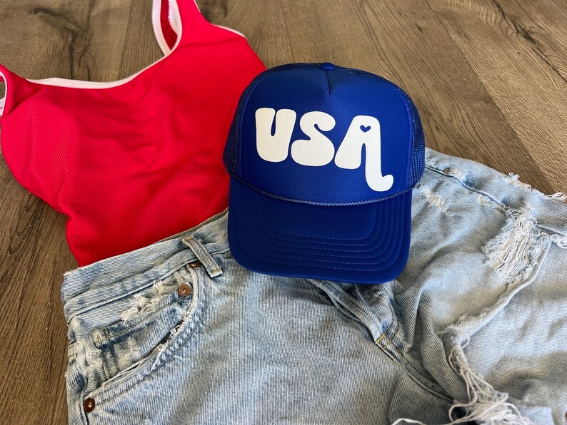 May include: A blue trucker hat with a white "USA" logo and a small heart. The hat is on top of a pair of light blue denim shorts and a red tank top.